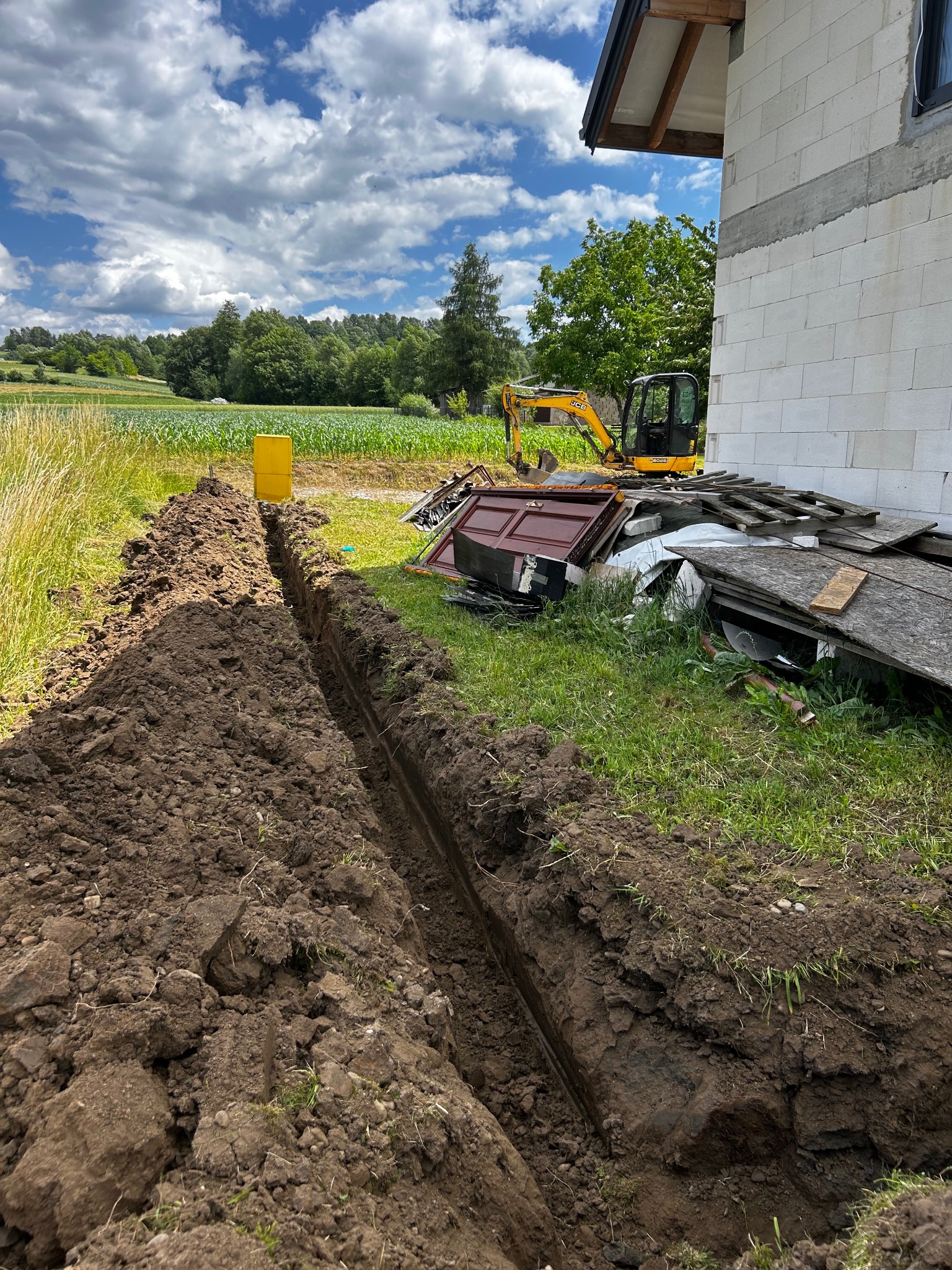 Wąski wykop w ziemi, z żółtym pojemnikiem i minikoparką w tle, obok budynku z białego bloczka. Widok na zielone pole i las.