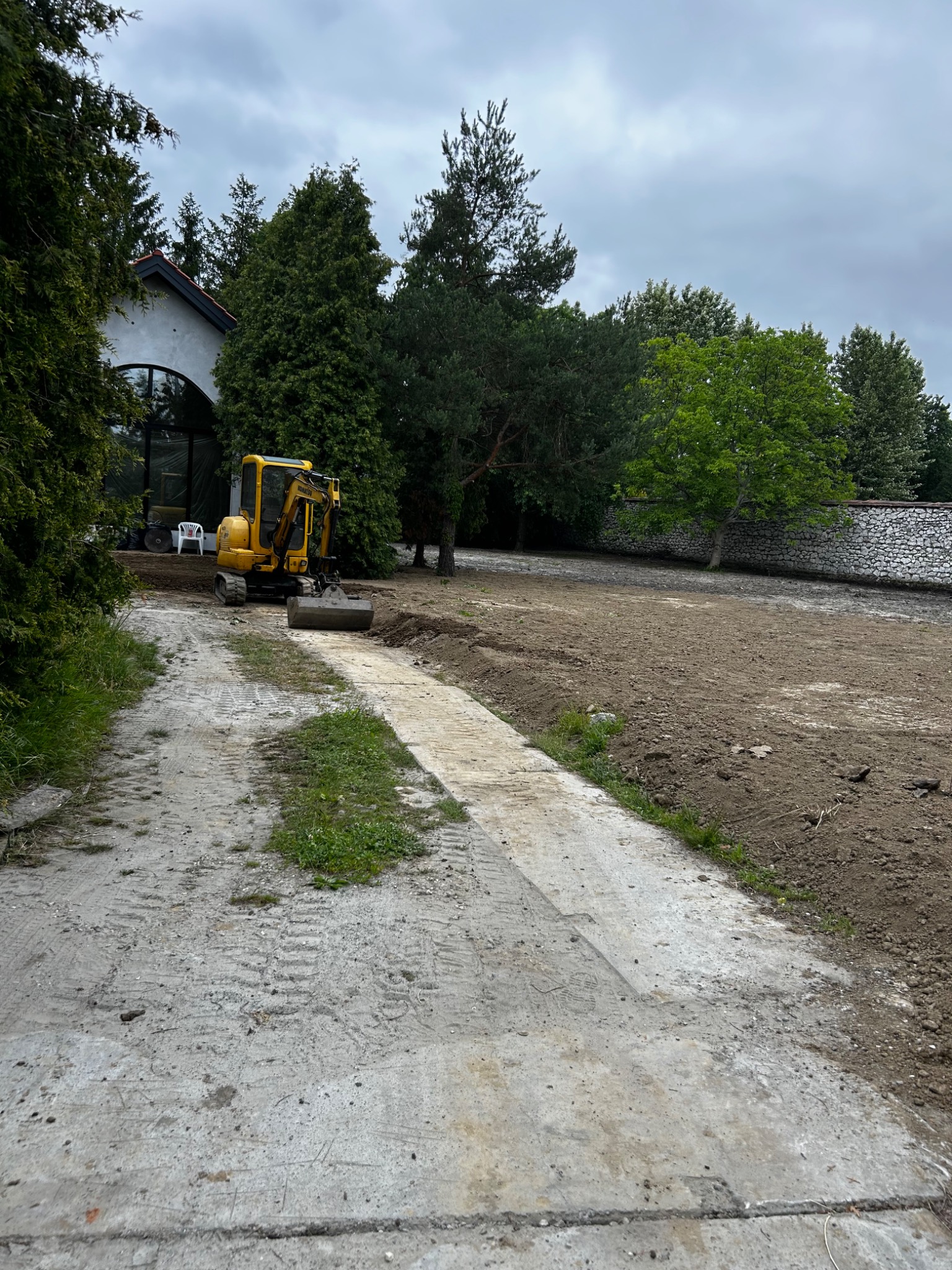 Żółta mini koparka na działce z przygotowanym terenem pod budowę, widoczny dom z dużym oknem łukowym i drzewa w tle.