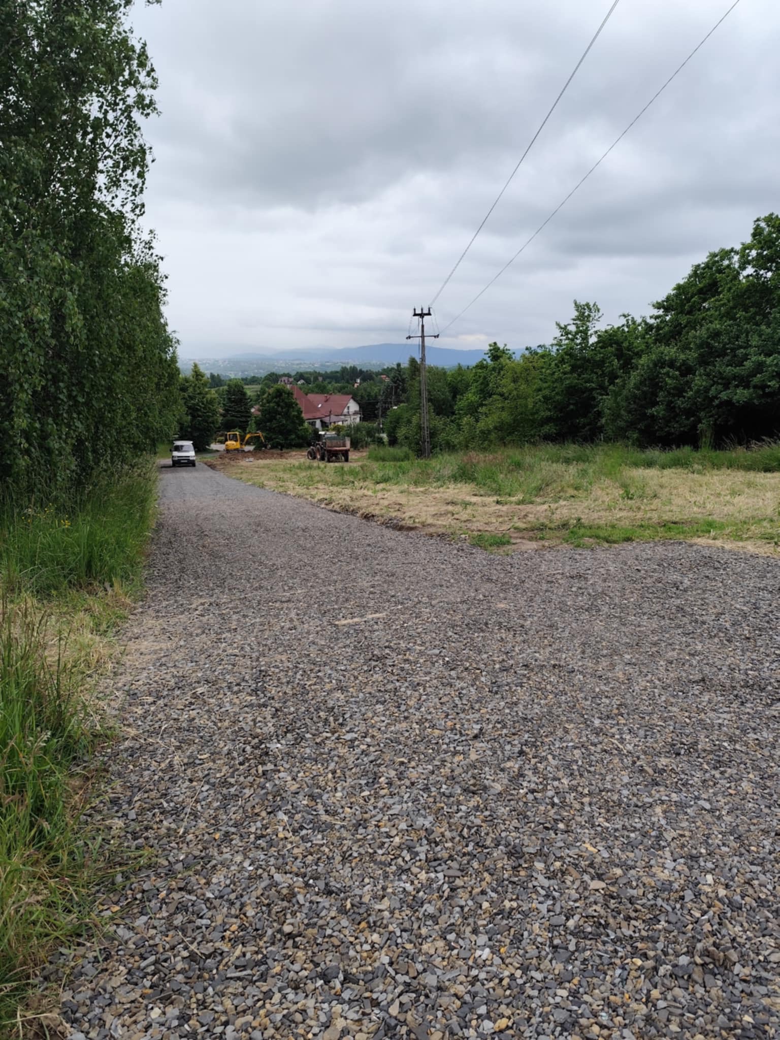 Utwardzona, szutrowa droga w perspektywie z oddalającym się białym busem i koparką w tle, otoczona zielenią i domami w oddali.