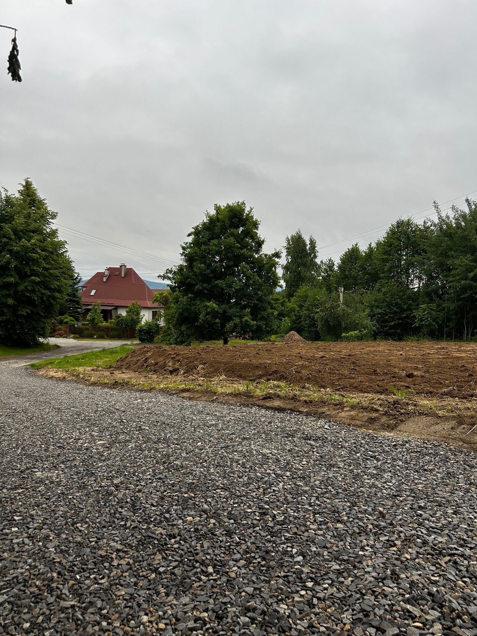 Przygotowany teren pod budowę z widoczną warstwą utwardzonego kruszywa i wyplantowaną ziemią, w tle dom z czerwoną dachówką i drzewa. Pochmurne niebo.