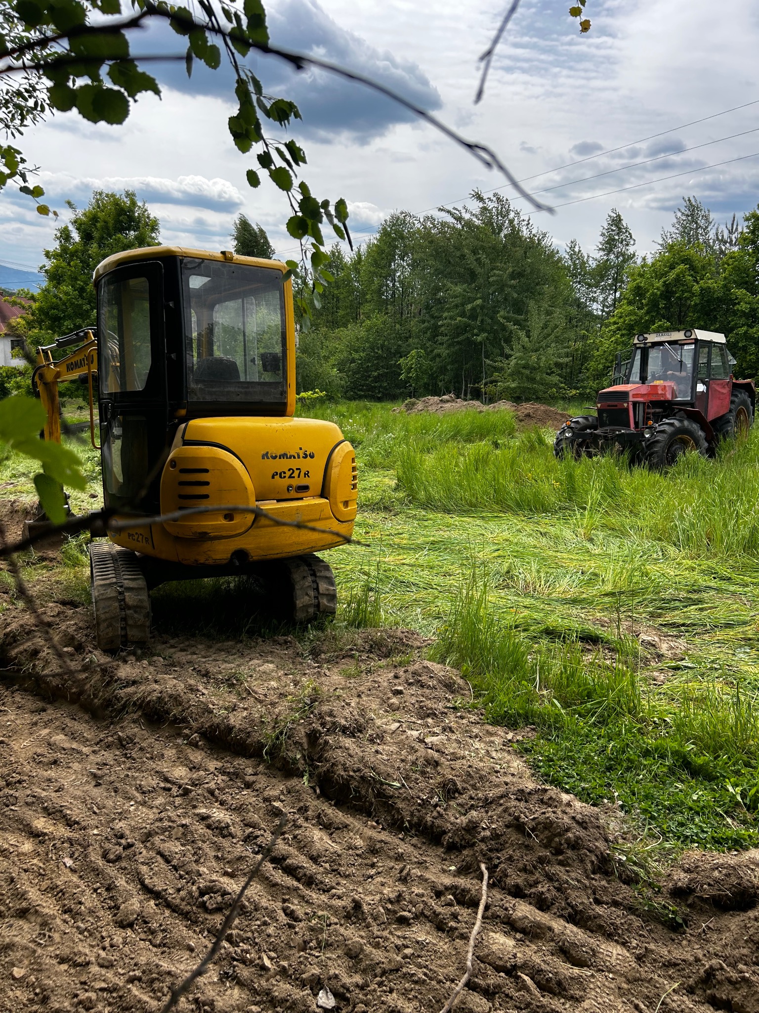 Żółta mini koparka Komatsu PC27R na nierównym terenie z widocznym śladem gąsienic i czerwony traktor Zetor w tle, otoczone zielenią i drzewami.