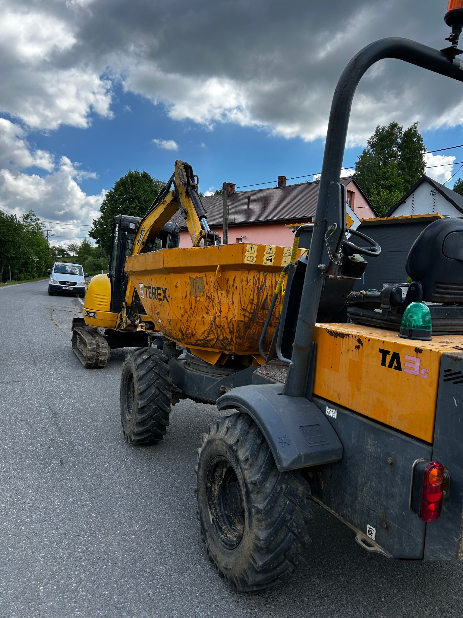Żółty mini-ładowarka Terex z łyżką, z załadowaną koparką gąsienicową na drodze asfaltowej, w tle budynki i biały samochód dostawczy. Praca na budowie.