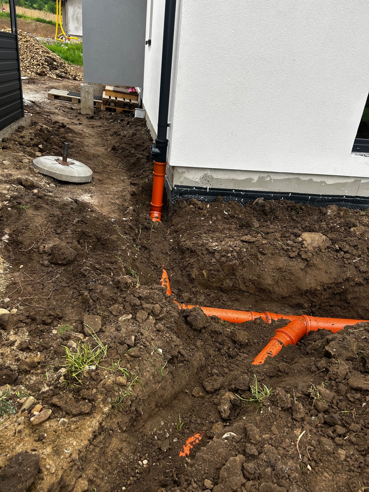 Pomarańczowe rury kanalizacyjne ułożone w wykopie ziemnym przy ścianie budynku z czarną rurą spustową. Widoczne palety i kamienie na podłożu.