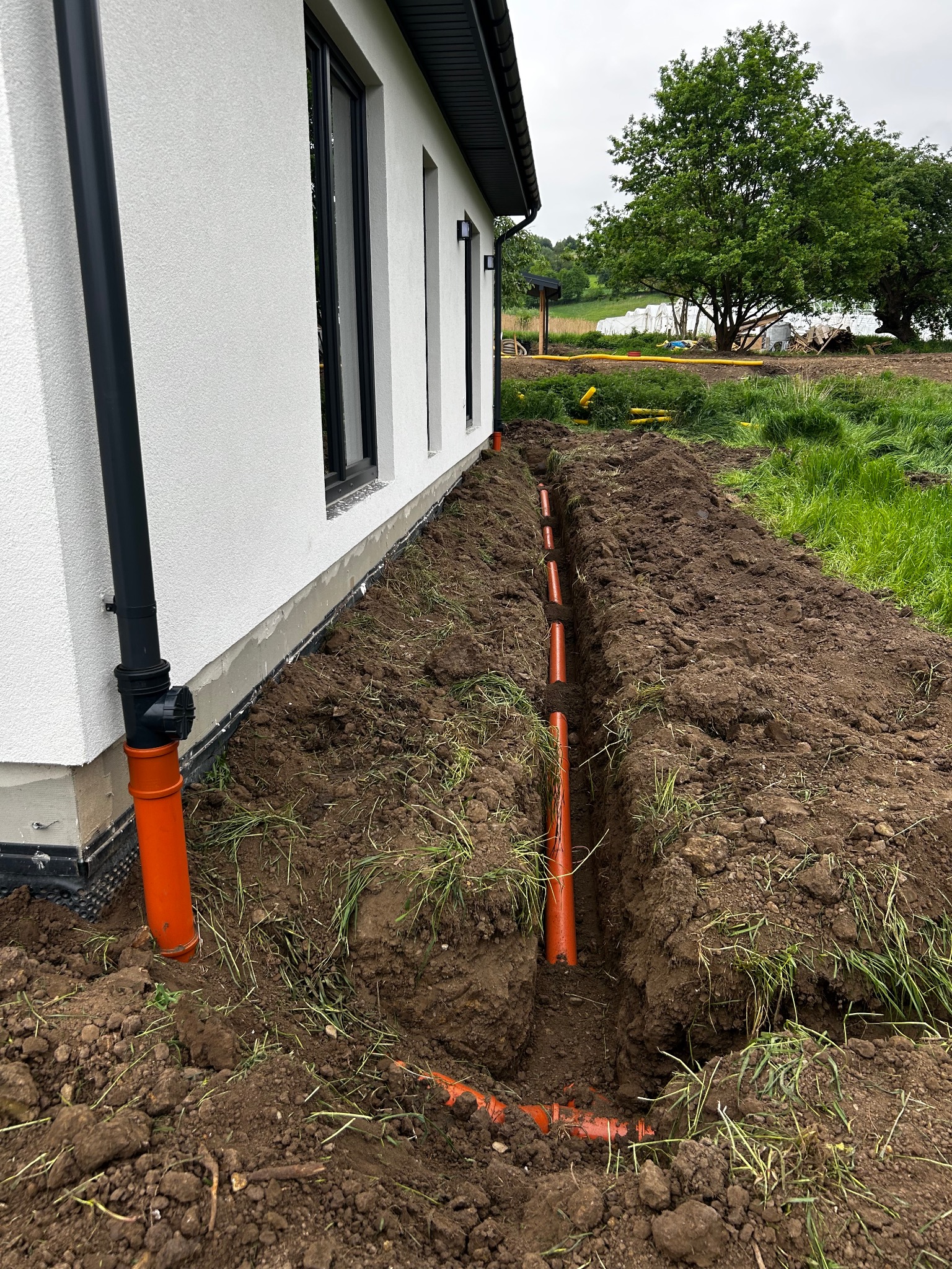 Pomarańczowe rury ułożone w wykopie wzdłuż białego budynku z czarnymi oknami. W tle trawa, drzewa i żółte rury. Widok z góry, lekko z boku.