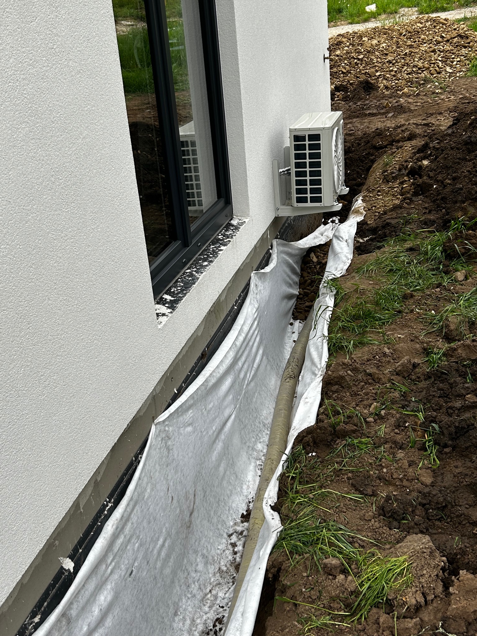 Wykop zabezpieczony geowłókniną wzdłuż ściany budynku z oknem i klimatyzatorem. Na dnie rura drenażowa, obok hałda kamieni.
