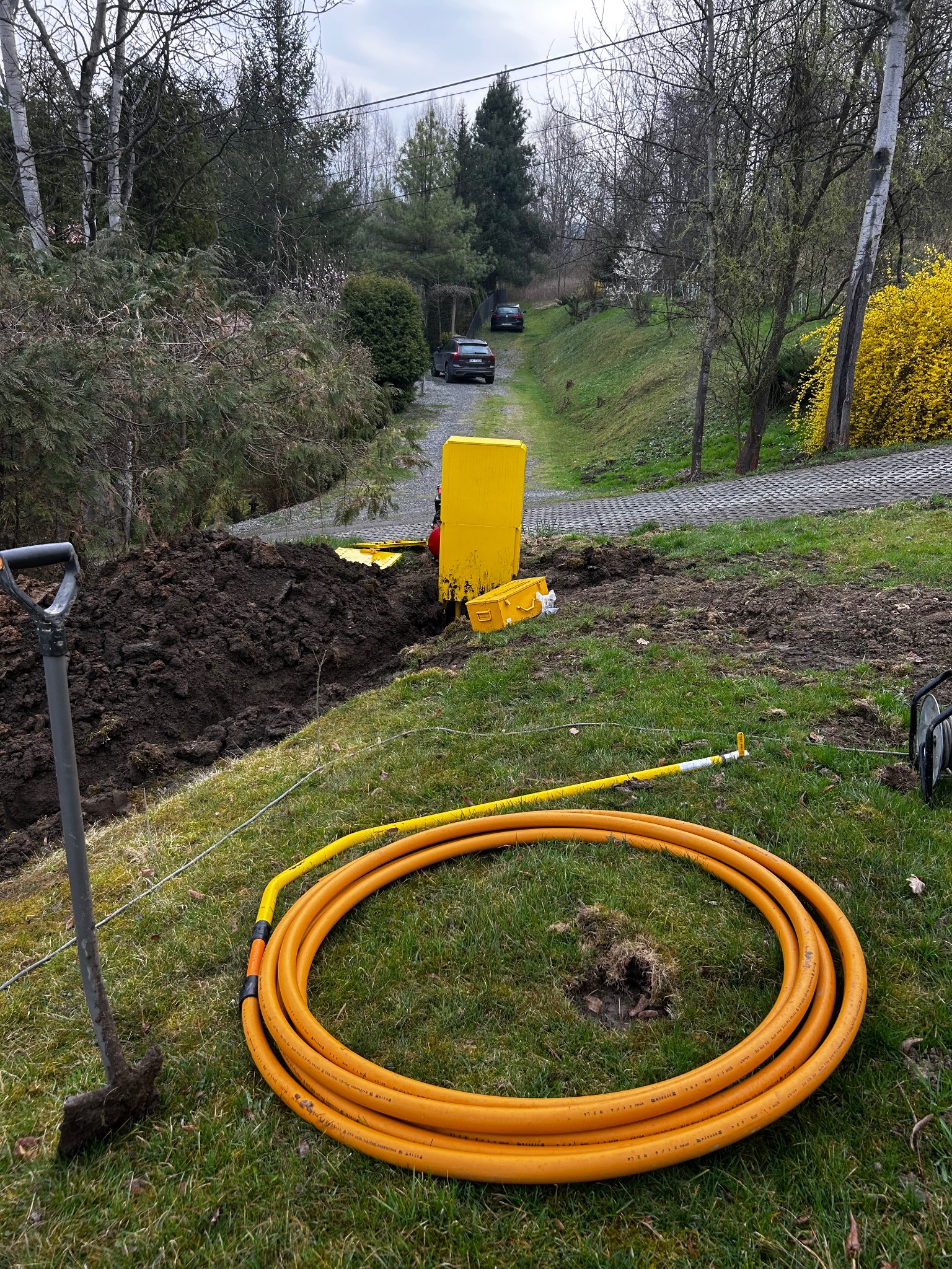 Żółty przewód gazowy zwinięty na trawie obok wykopu z łopatą i skrzynką narzędziową na tle zieleni i podjazdu z samochodami.