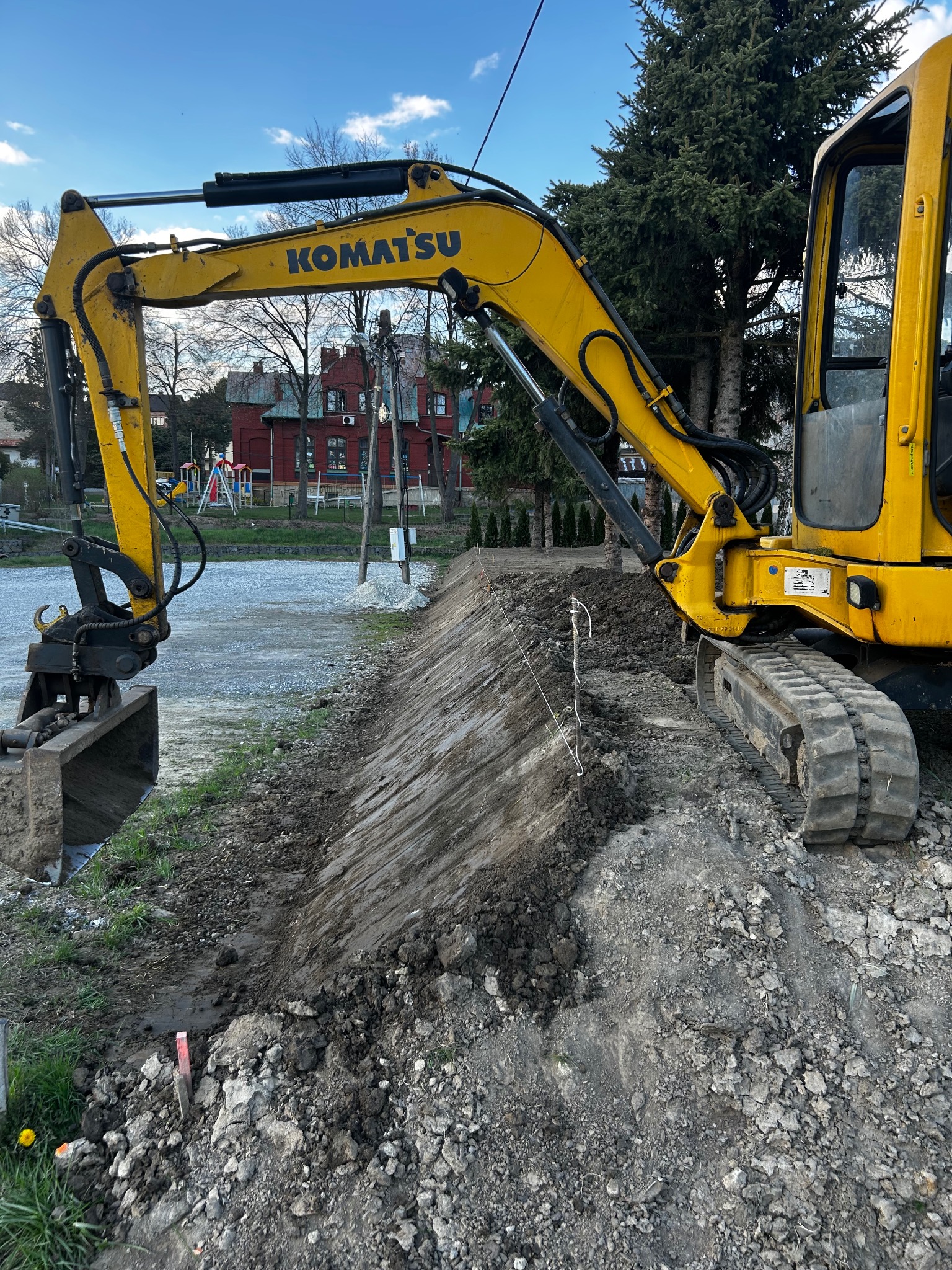 Żółta koparka Komatsu na placu budowy, wyrównany teren z widocznymi śladami pracy łyżki, w tle budynek z czerwonej cegły i plac zabaw.