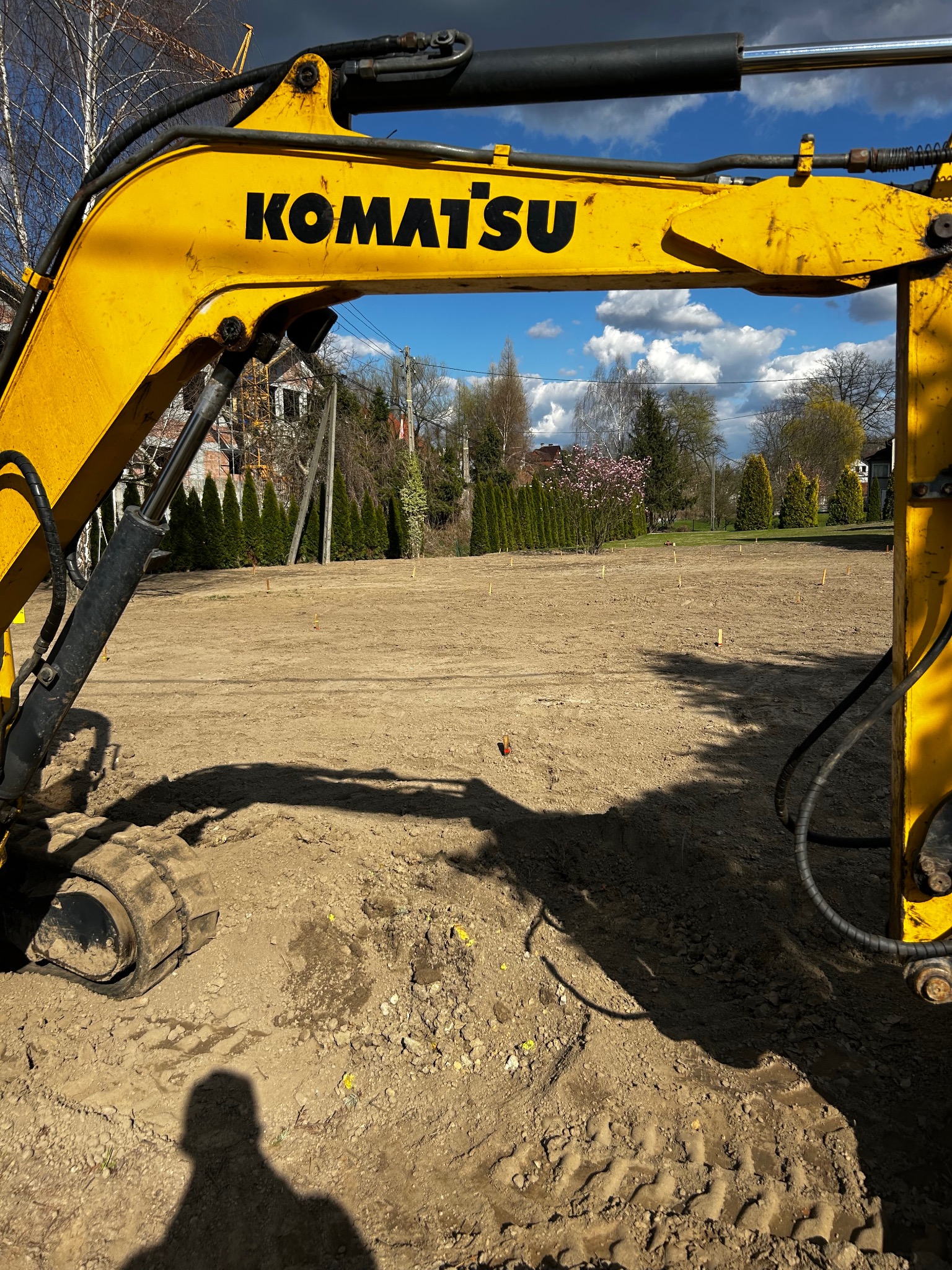 Żółta koparka Komatsu na placu budowy z wbitymi palikami, w tle domy i błękitne niebo z chmurami. Widoczny cień osoby.