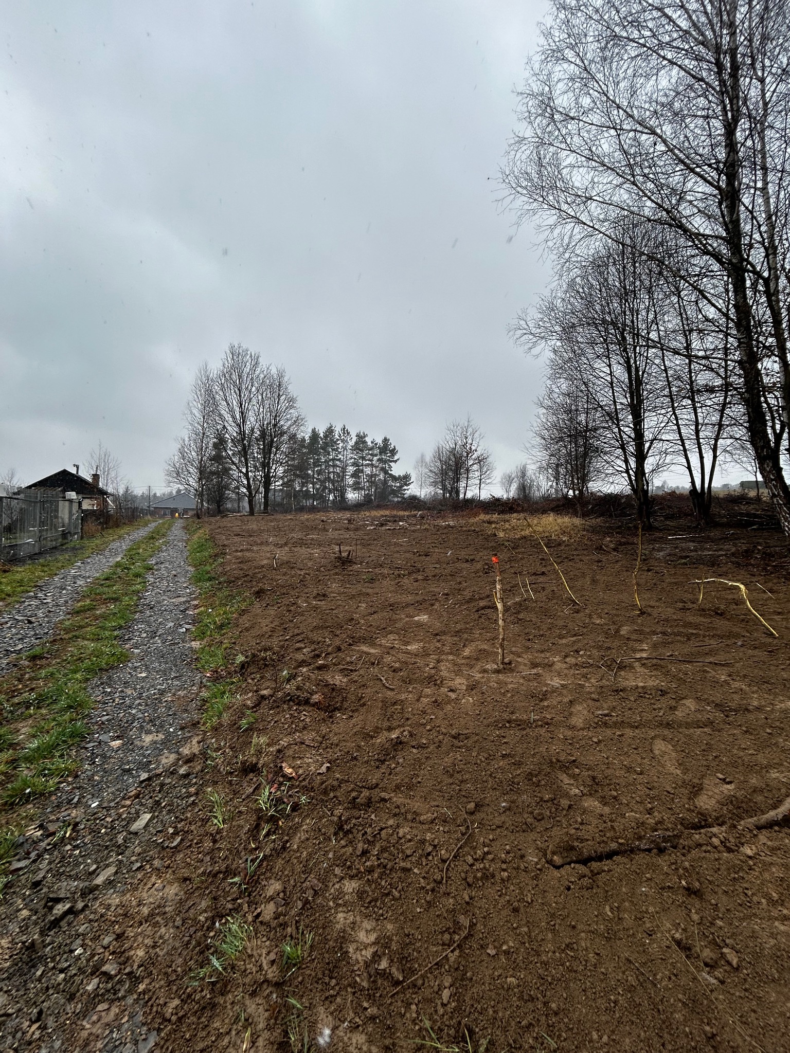 Przygotowany teren pod budowę w pochmurny dzień. Widoczne wytyczone granice, świeżo przekopana ziemia i ślady po wycince drzew. W tle budynki i drzewa.