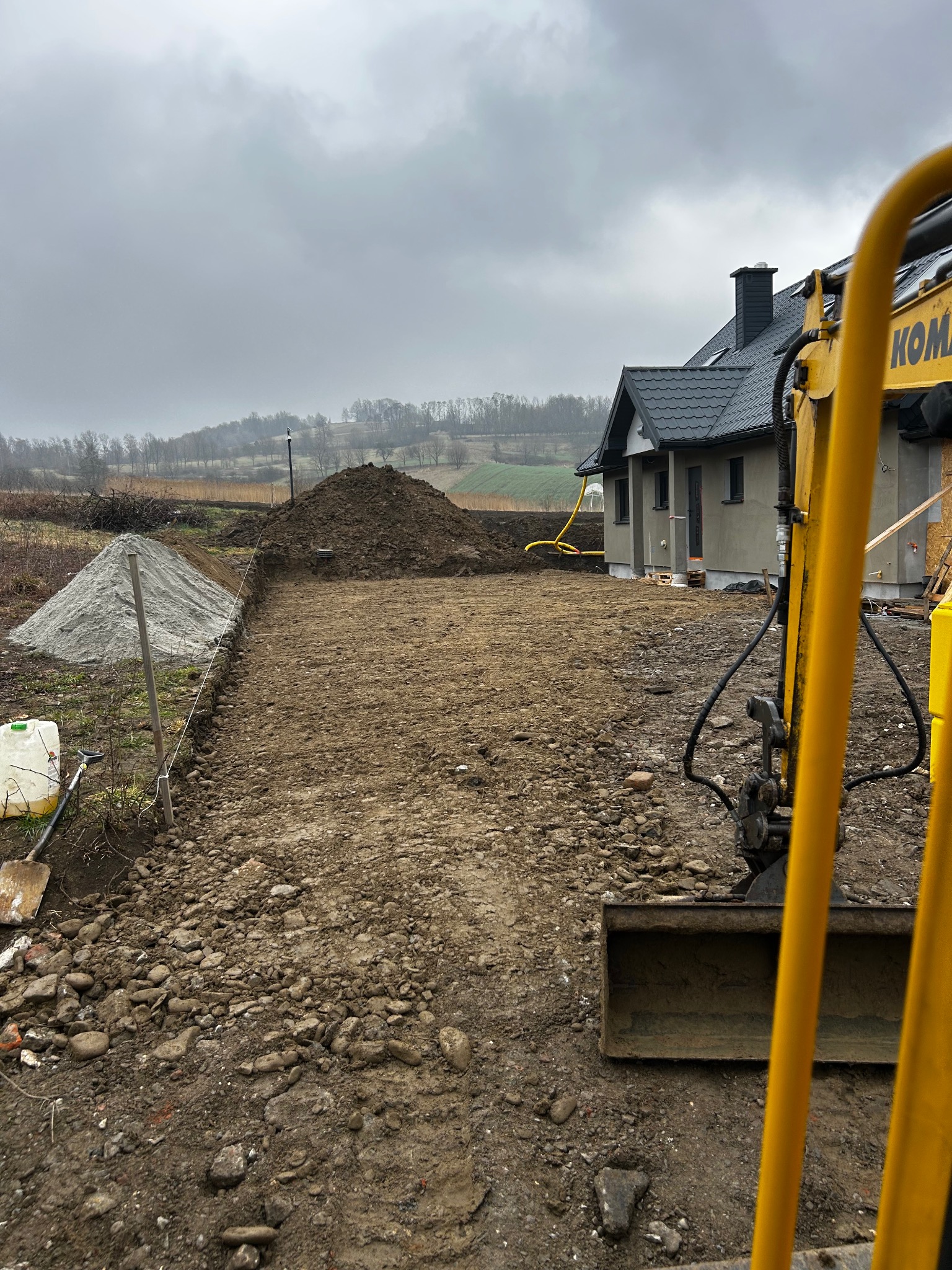 Przygotowany teren pod budowę domu jednorodzinnego z widocznym wykopem, stertą ziemi i piasku, oraz fragmentem koparki na pierwszym planie w pochmurny dzień.