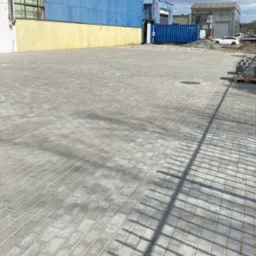 Szara, ukończona nawierzchnia z kostki brukowej na posesji. W tle budynki przemysłowe, zaparkowany samochód i paleta z kostką przy metalowym ogrodzeniu.