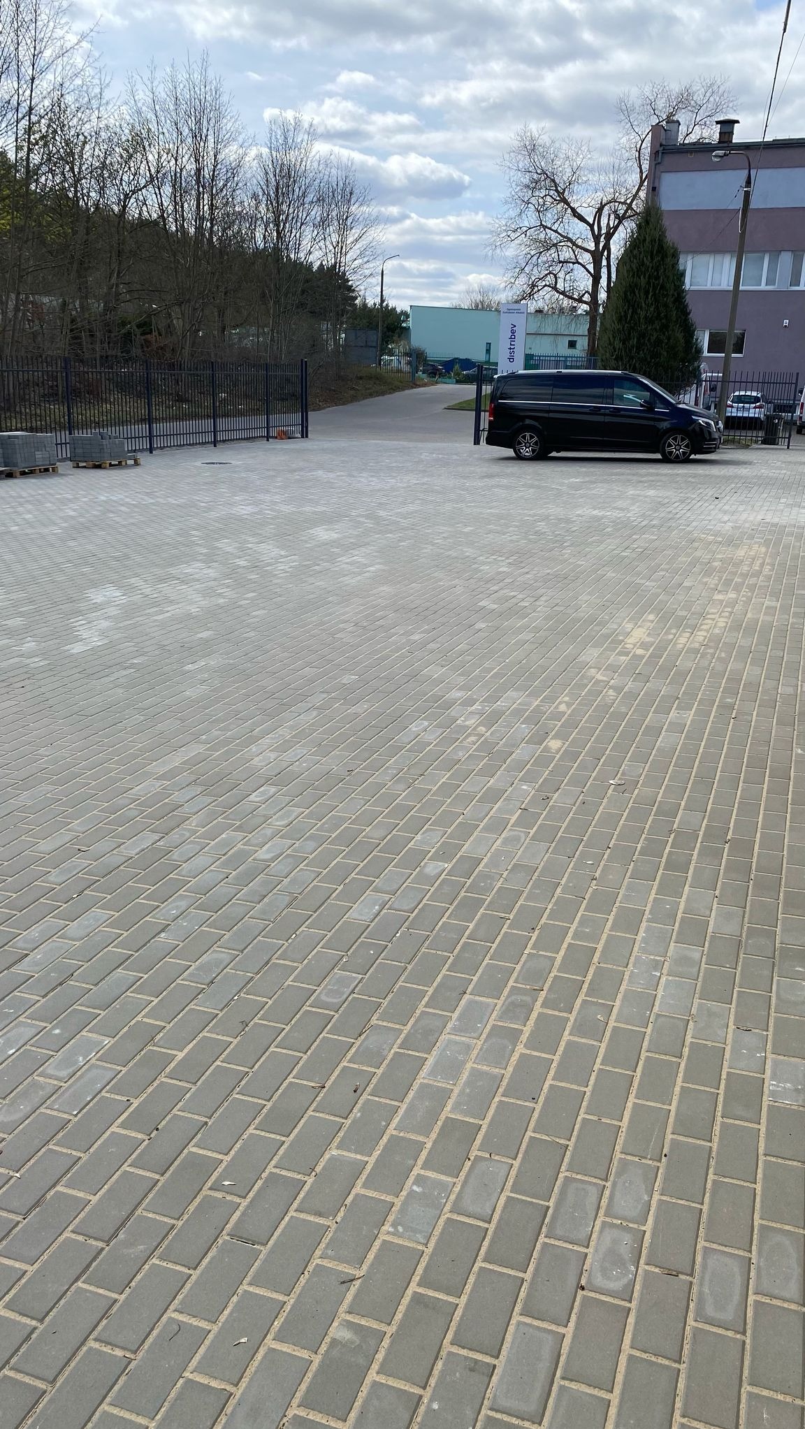 Szara, prostokątna kostka brukowa na posesji firmy, z czarnym vanem w tle, brama wjazdowa i budynek. Regularny wzór ułożenia kostki. Widok z perspektywy.