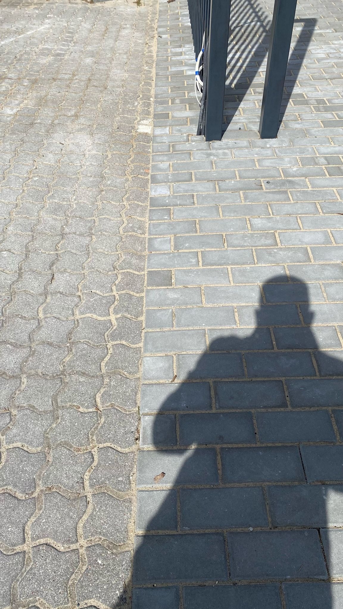 Porównanie kostki: stara, szara i nowa, grafitowa, ułożona w cegiełkę. Cień osoby na nowej kostce. Elementy bramy widoczne w prawym górnym rogu.