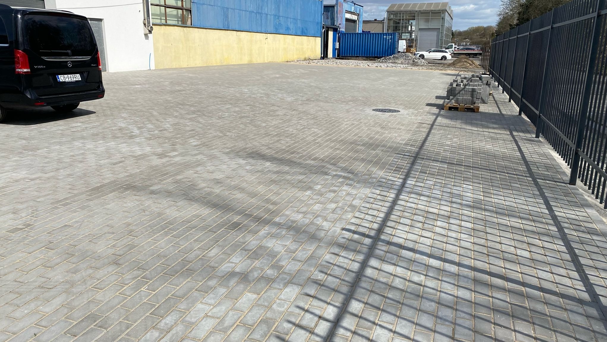 Szara, ukończona nawierzchnia z kostki brukowej na posesji. W tle budynki przemysłowe, zaparkowany samochód i paleta z kostką przy metalowym ogrodzeniu.