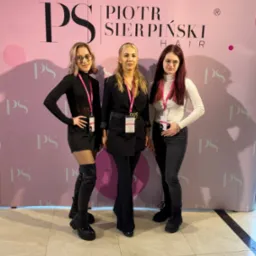 Trzy kobiety pozują na tle ścianki reklamowej Piotr Sierpiński Hair, prezentując różne stylizacje włosów i ubrania, z identyfikatorami na szyjach.