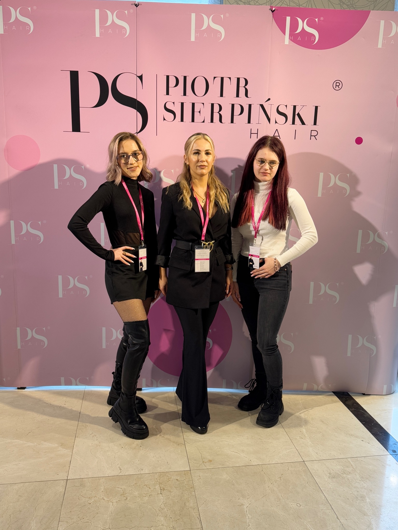 Trzy kobiety pozują na tle ścianki reklamowej Piotr Sierpiński Hair, prezentując różne stylizacje włosów i ubrania, z identyfikatorami na szyjach.