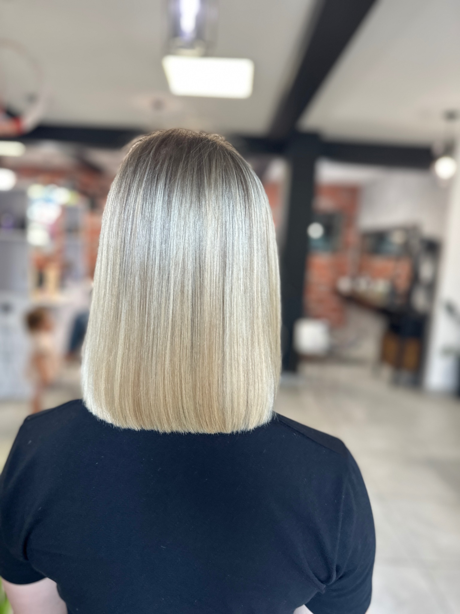 Tył głowy kobiety z prostymi, blond włosami ombre, świeżo po strzyżeniu na boba, widoczne ciemniejsze odrosty i jasne końcówki, w tle salon fryzjerski z ceglaną ścianą.