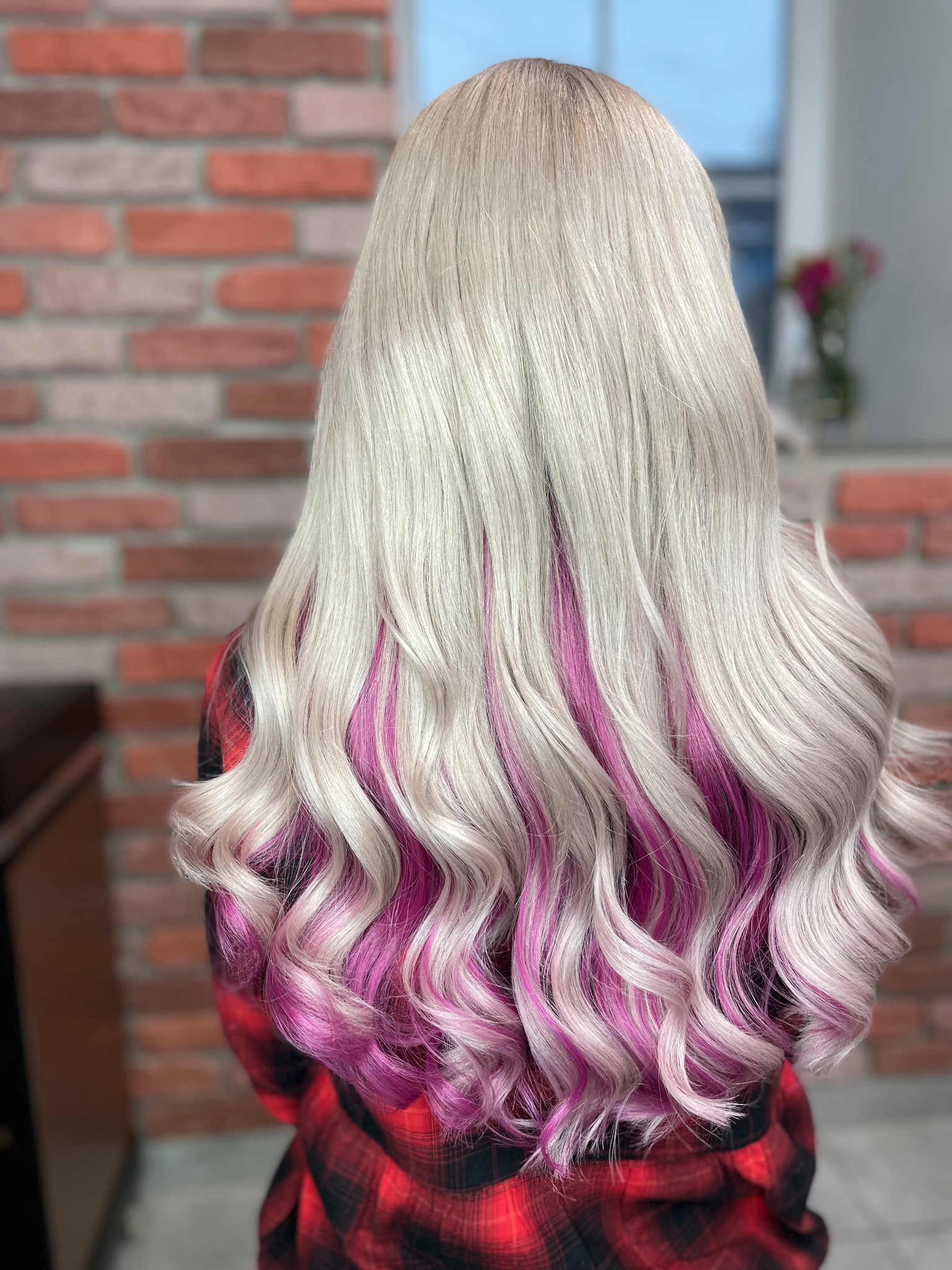 Długie, platynowe włosy z różowymi pasemkami u nasady, ułożone w loki, widok z tyłu na tle ceglanej ściany.