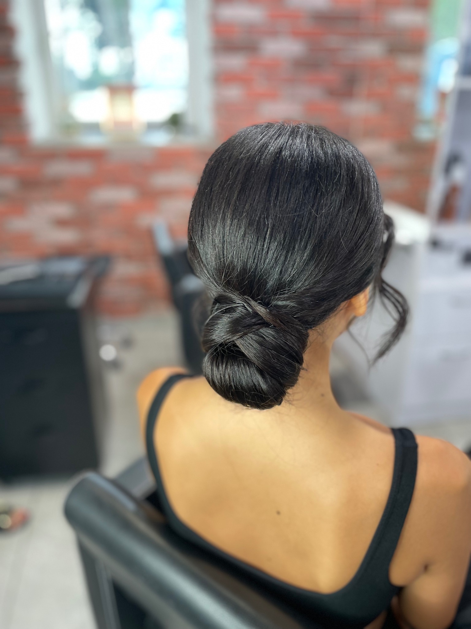 Eleganckie upięcie włosów w kok hiszpański, widok z tyłu, z pojedynczymi kosmykami swobodnie opadającymi na kark, na tle ściany z czerwonej cegły w salonie.