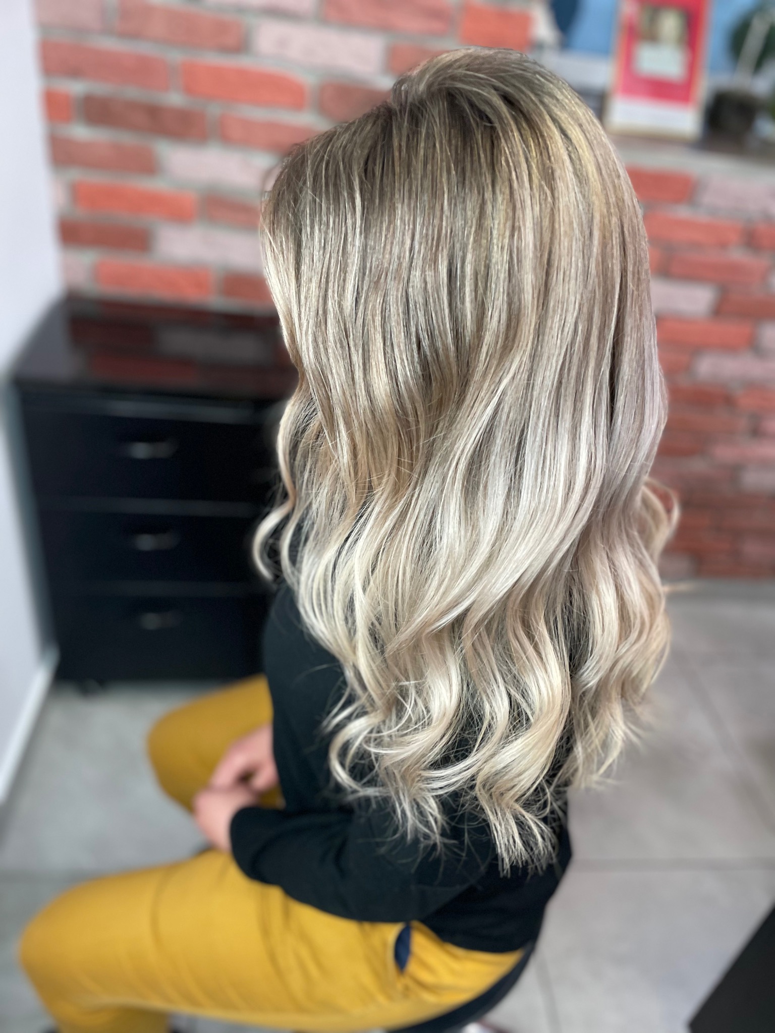 Długie, falowane włosy z balejażem w odcieniach blondu i beżu, widoczne z tyłu, na tle ceglanej ściany i czarnej komody.