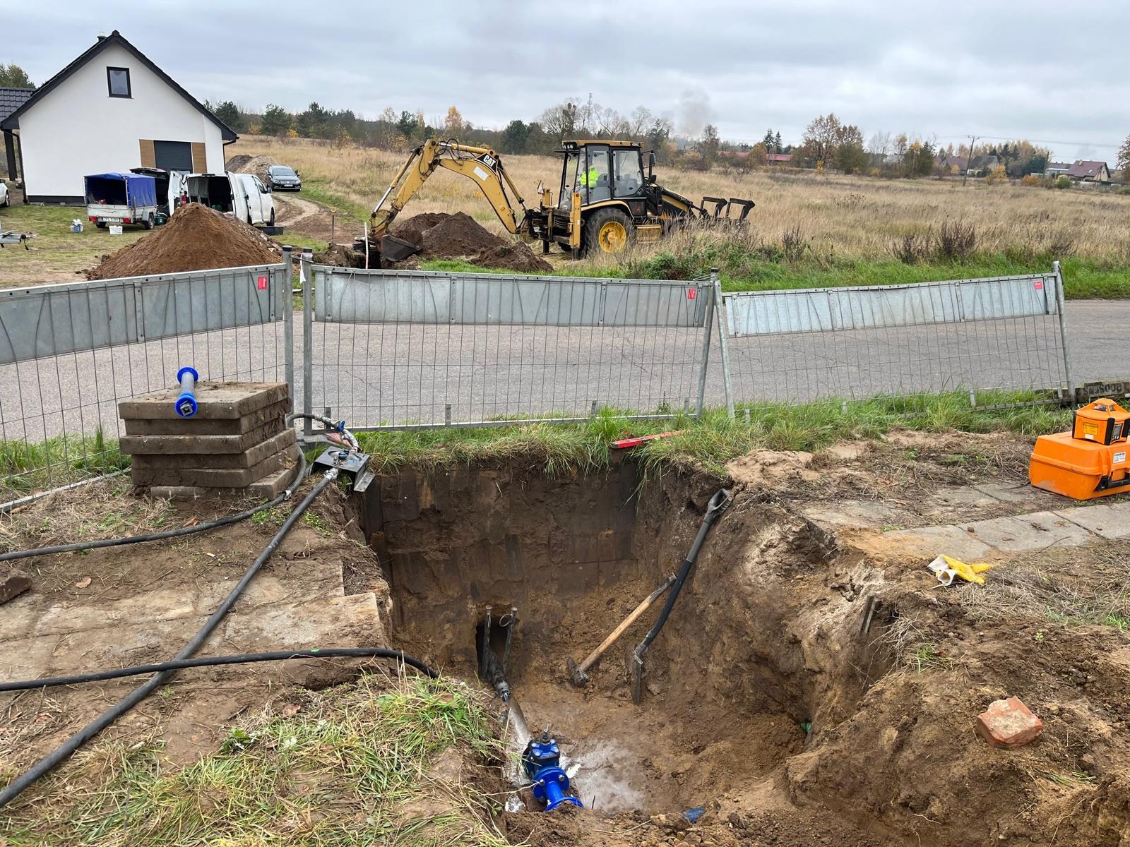 Wykop z widocznymi rurami i niebieskim zaworem, prace ziemne wykonywane koparką CAT, ogrodzenie tymczasowe, zagęszczarka wibracyjna, teren budowy.