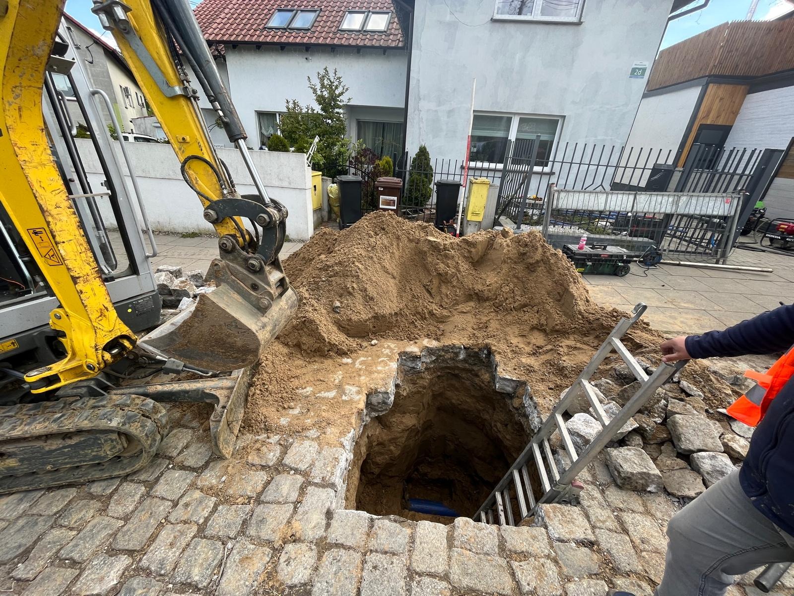 Wykop w brukowanej ulicy z żółtą koparką obok, widoczna drabina wewnątrz wykopu i fragment niebieskiej rury na dnie, obok sterta piasku.