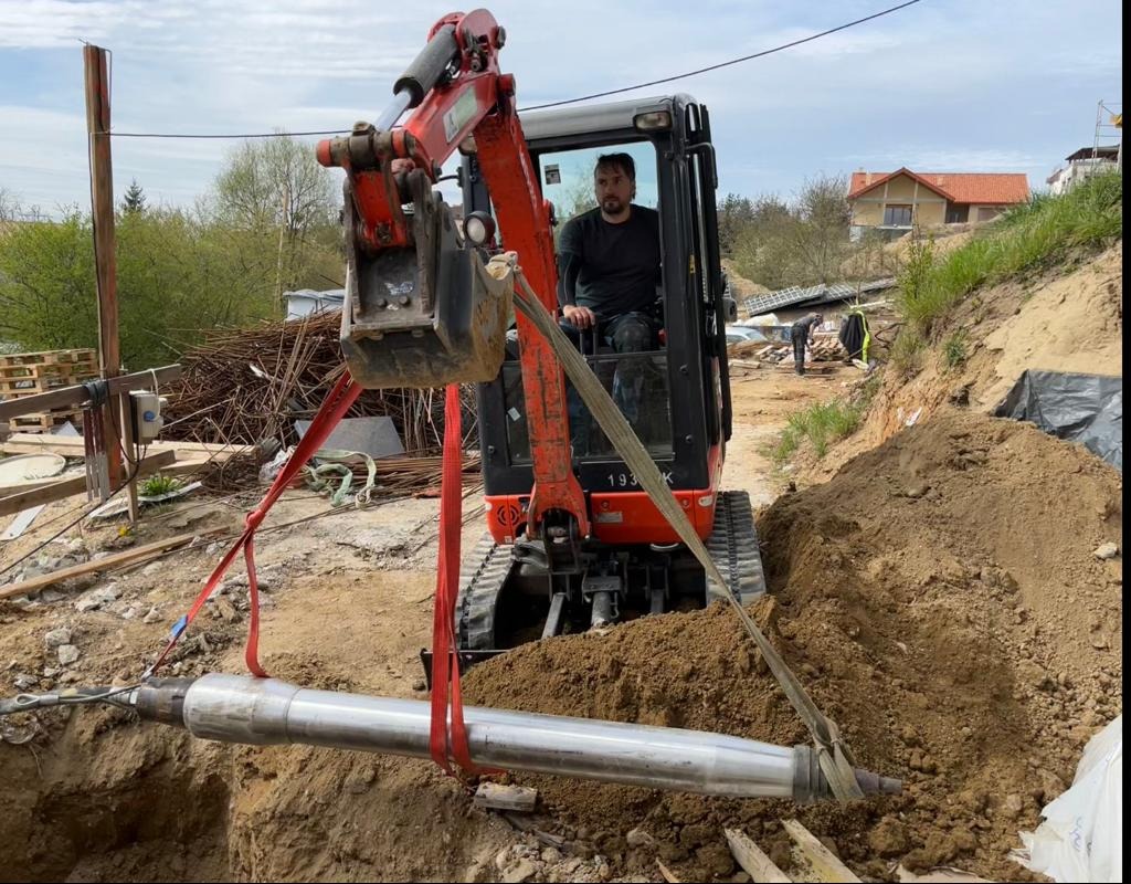 Operator w minikoparce opuszcza rurę do wykopu za pomocą pasów transportowych, widoczne zbrojenia i palety w tle.