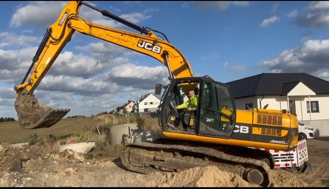 Żółta koparka JCB podczas prac ziemnych na budowie osiedla domów jednorodzinnych, operator w kabinie w kamizelce odblaskowej, widoczne betonowe kręgi.