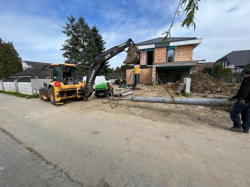 Żółta koparko-ładowarka Volvo BL71B podnosi rurę za pomocą łyżki, w tle budynek w stanie surowym i dwaj mężczyźni przyłączeni do rury.