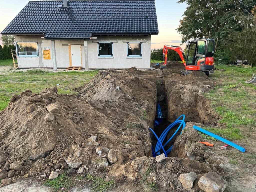 Wykonanie wykopu pod przyłącze wodociągowe z widocznymi niebieskimi rurami w wykopie, mała koparka i budynek w stanie surowym otwartym w tle.