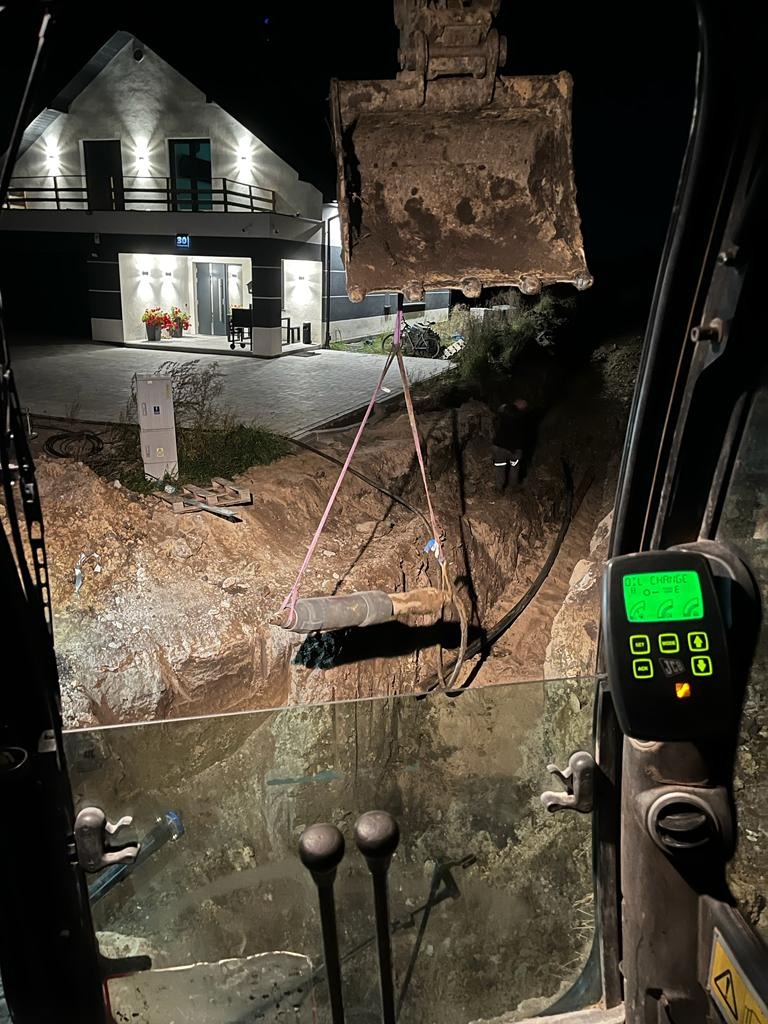 Widok z kabiny koparki na wykop w ziemi, z zawieszonym elementem instalacji za pomocą pasów, w tle oświetlony budynek mieszkalny nocą.