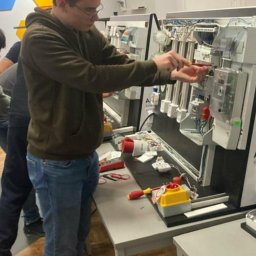 BTT-electrics. Usługi Elektryczne Arkadiusz Najmrodzki - Młody elektryk w okularach pracuje przy panelu z przewodami i miernikami, używając śrubokręta. Na stole leżą narzędzia i elementy instalacji.