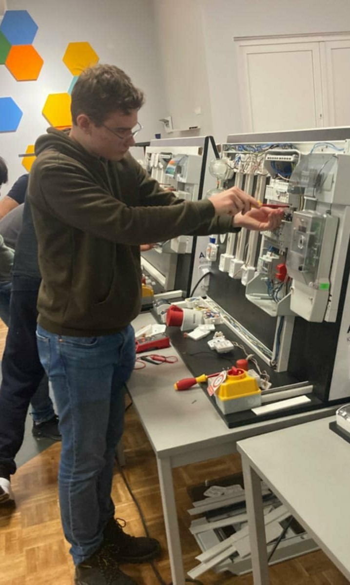 Młody elektryk w okularach pracuje przy panelu z przewodami i miernikami, używając śrubokręta. Na stole leżą narzędzia i elementy instalacji.