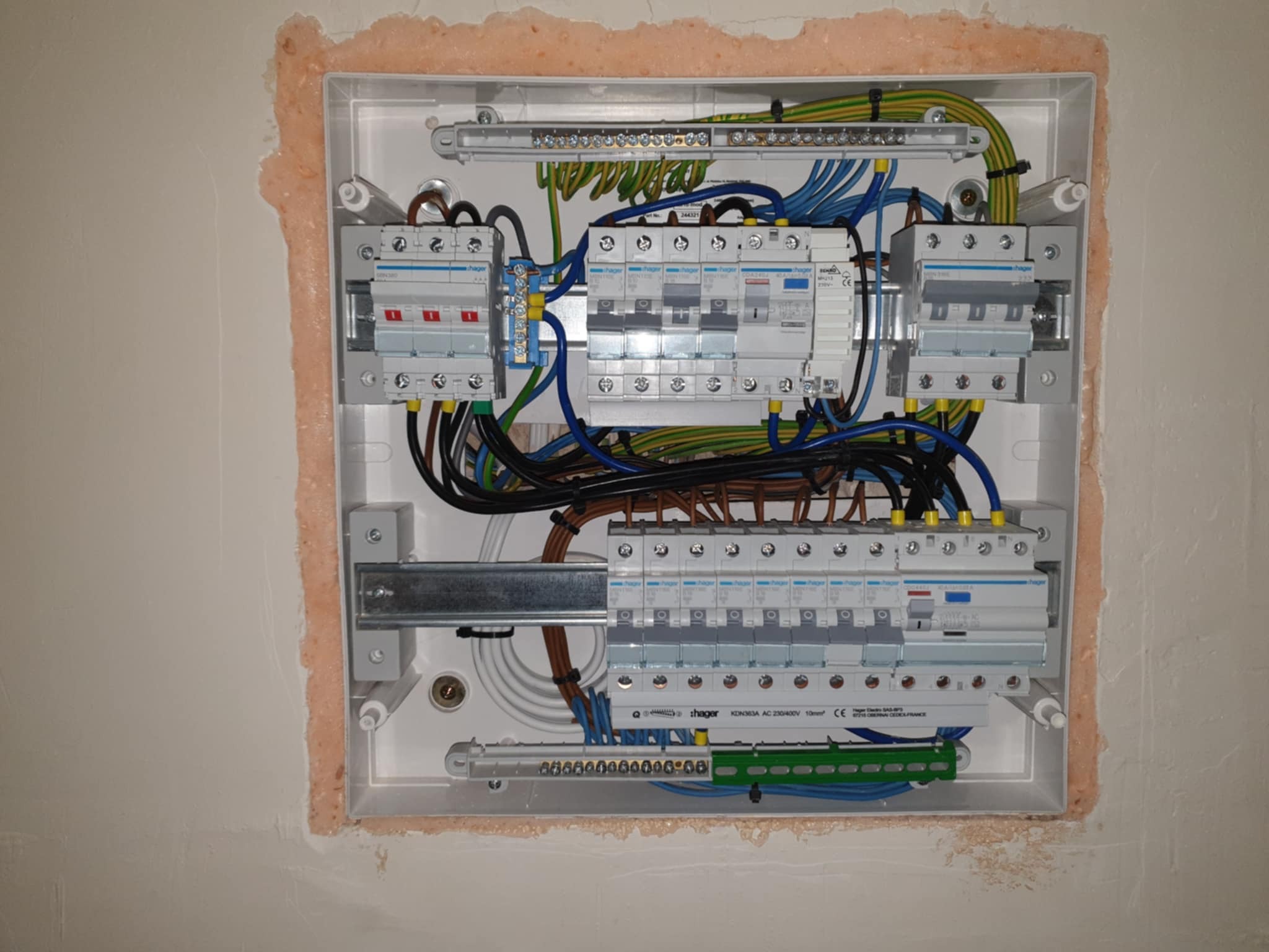 Rozdzielnica elektryczna Hager w białej obudowie, zamontowana w ścianie z widocznymi przewodami i zabezpieczeniami. Widoczne ślady po montażu wokół obudowy.