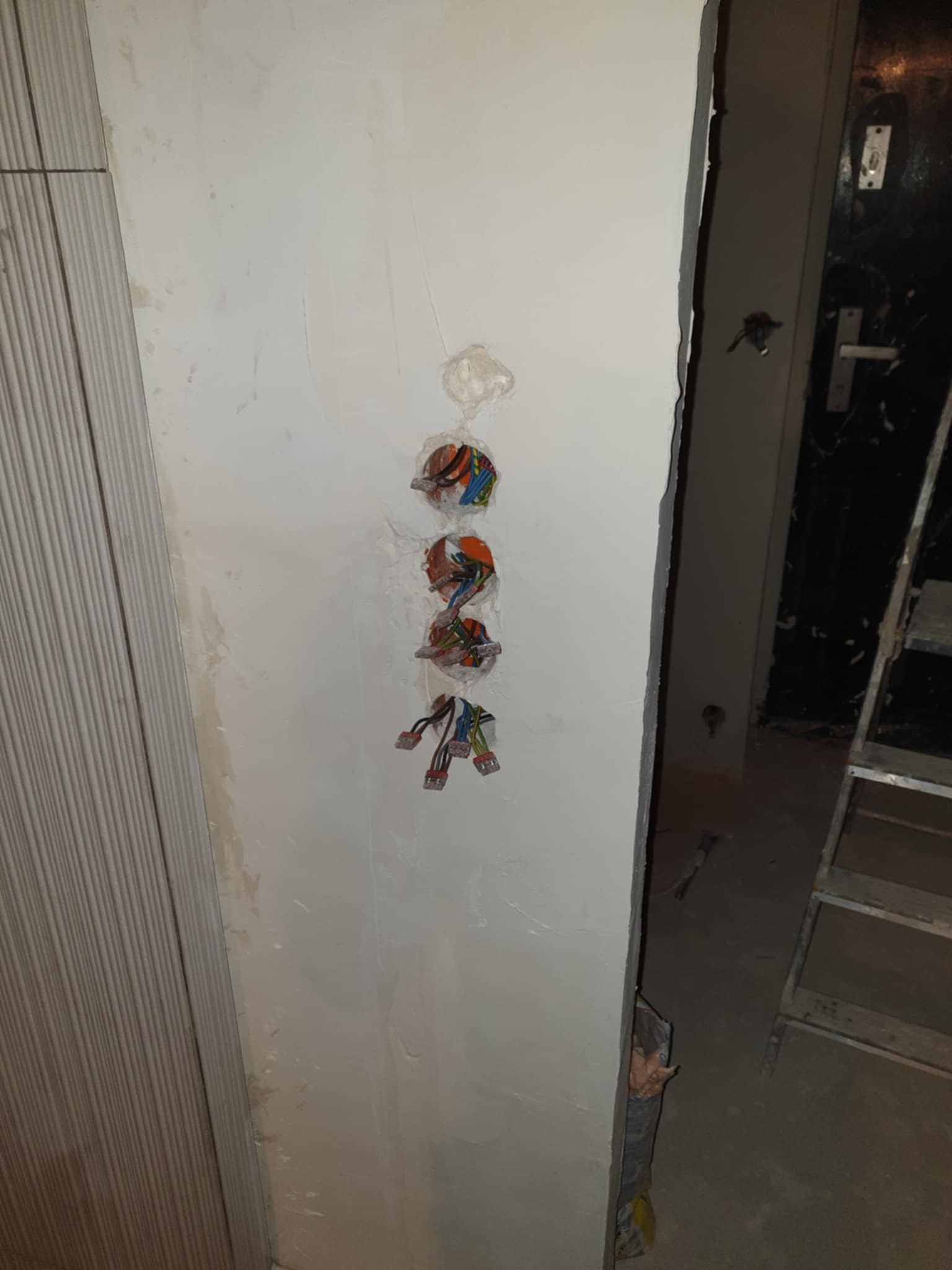 Ściana z otworami pod gniazdka elektryczne i wystającymi przewodami w kolorowej izolacji. Widoczny fragment drabiny i drzwi w tle. Prace instalacyjne w trakcie.