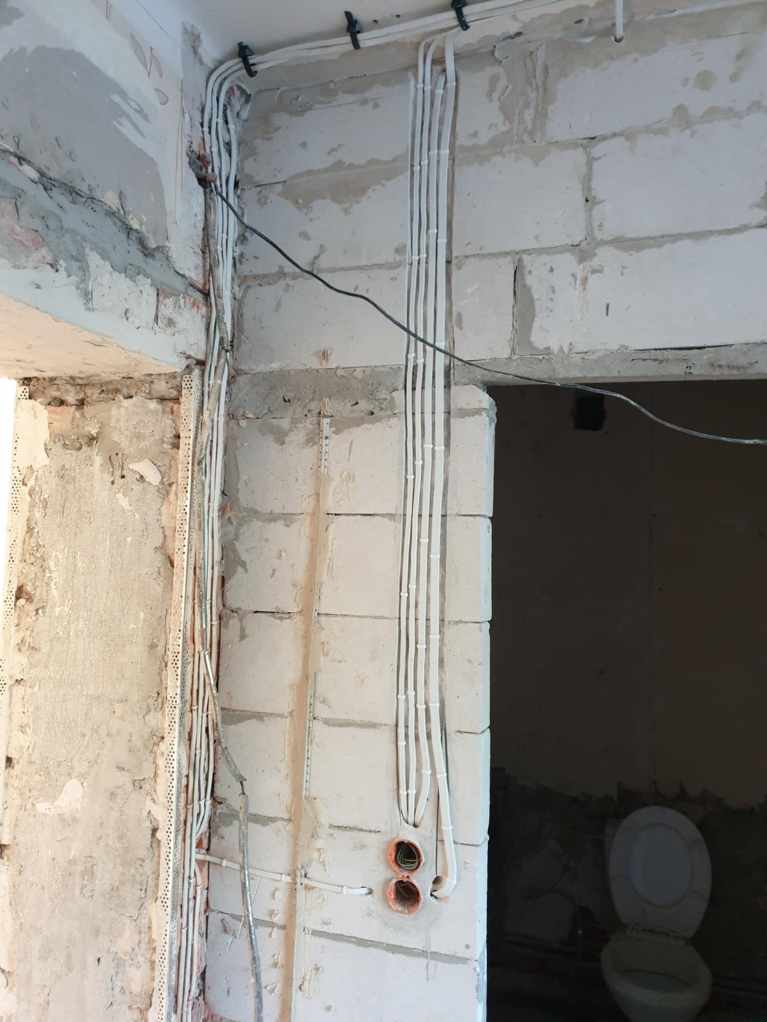 Białe kable elektryczne równolegle ułożone na surowej ścianie z betonu komórkowego w trakcie remontu, widoczne puszki instalacyjne i uchwyty mocujące.