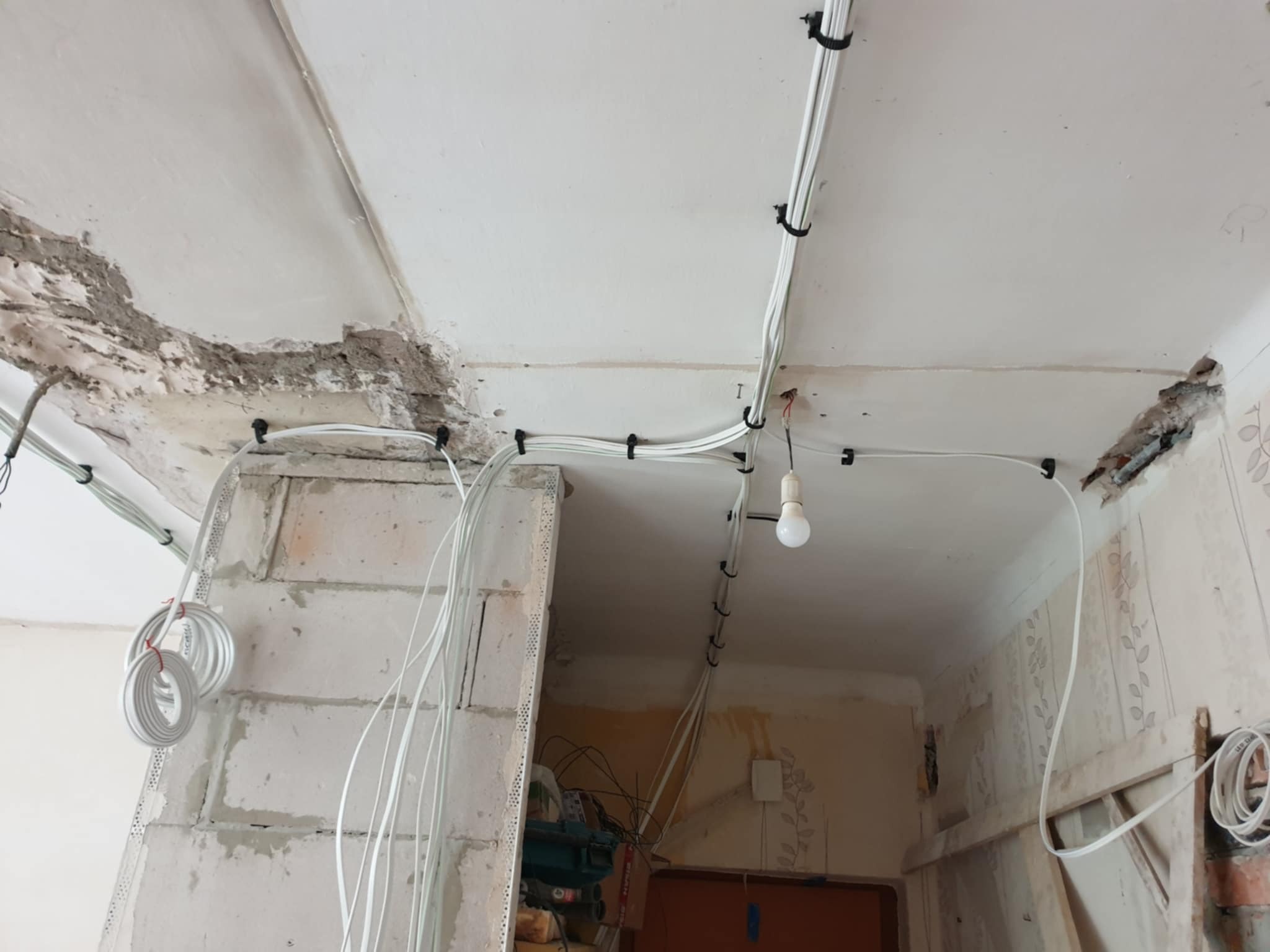 Białe przewody elektryczne przymocowane do sufitu i ściany za pomocą czarnych uchwytów. Widoczna surowa ściana i prowizoryczna żarówka.