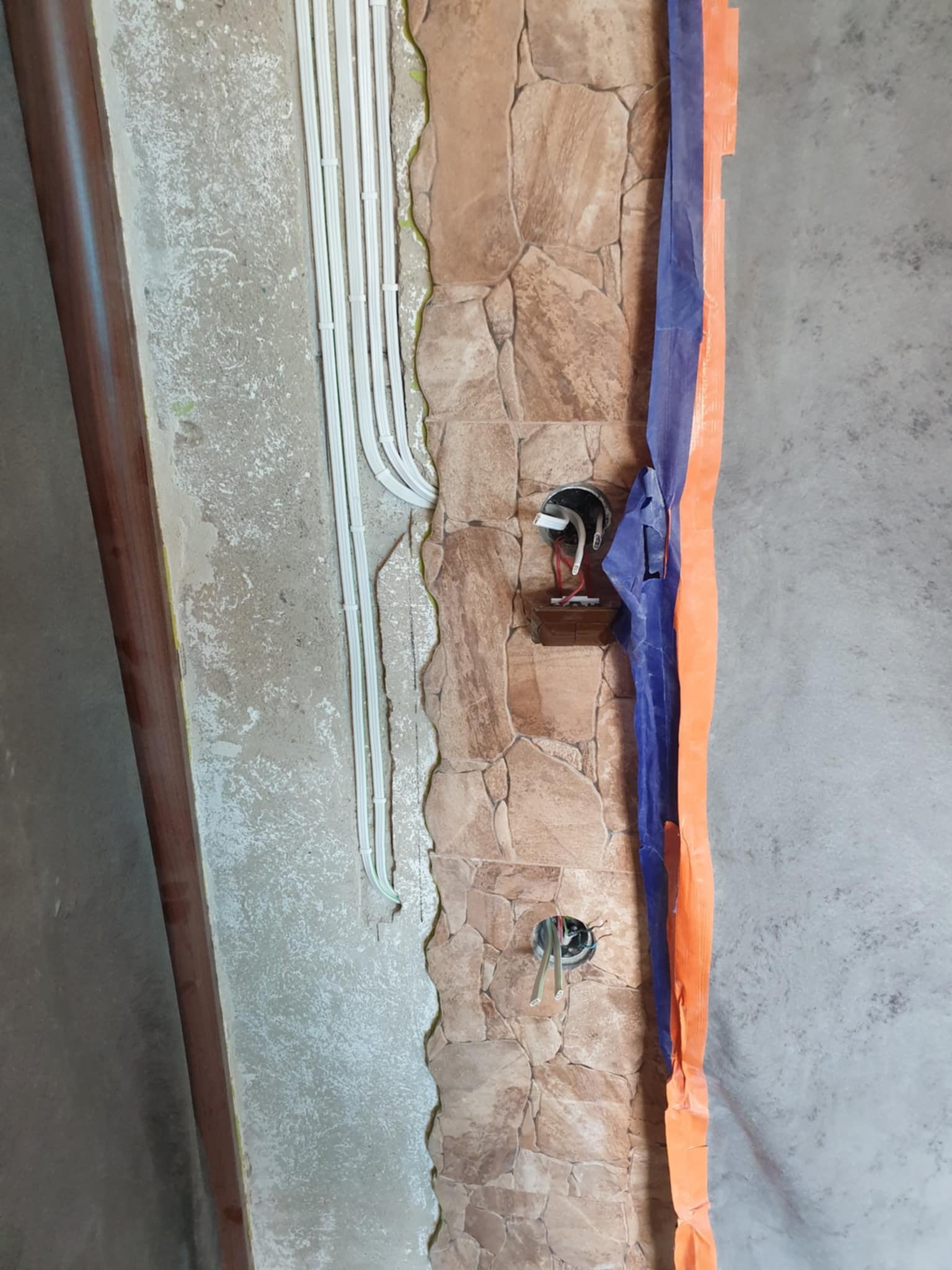 Montaż instalacji elektrycznej w ścianie z imitacją kamienia, widoczne kable w peszlach i puszki instalacyjne z przewodami, zabezpieczone folią malarską.