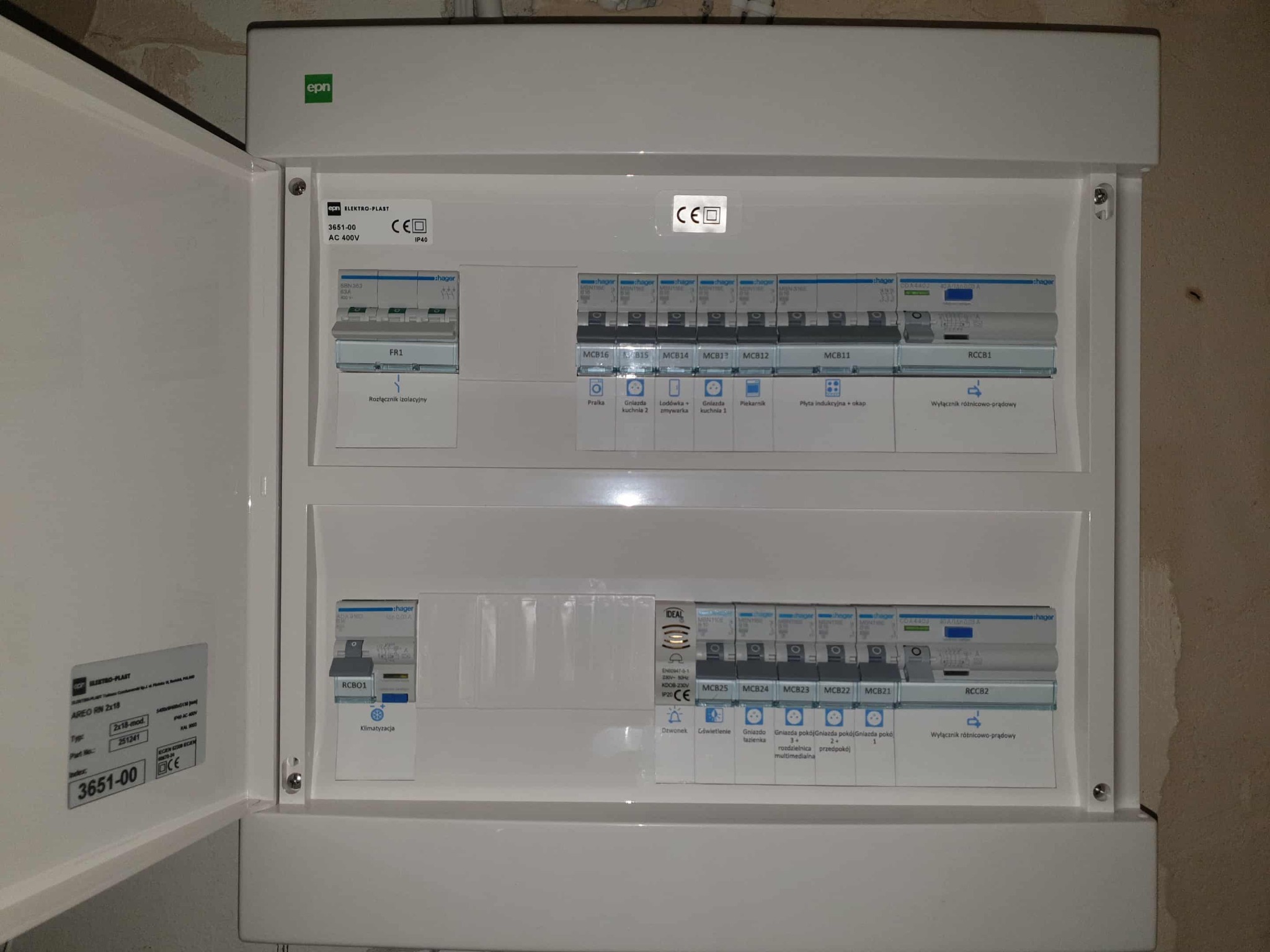 Otwarta, biała skrzynka rozdzielcza z bezpiecznikami i oznaczeniami, widoczne elementy instalacji elektrycznej wewnątrz. Detal: etykiety z opisem obwodów.