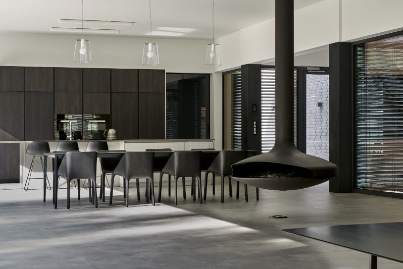 Nowoczesna jadalnia z ciemnymi meblami, wiszącym kominkiem i betonową podłogą, oświetlona przez minimalistyczne lampy.