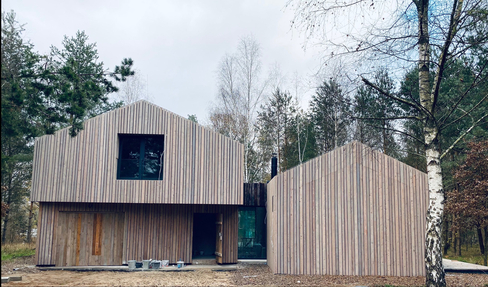 Nowoczesny dom z drewnianą elewacją w skandynawskim stylu, widoczne puszki z farbą na zewnątrz, otoczony lasem.