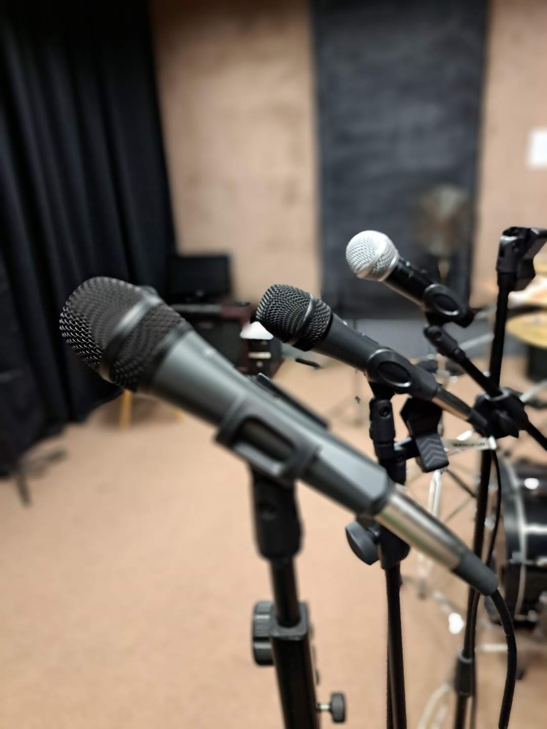 Trzy mikrofony na statywach w studiu nagraniowym, z perkusją w tle i czarną zasłoną po lewej stronie.