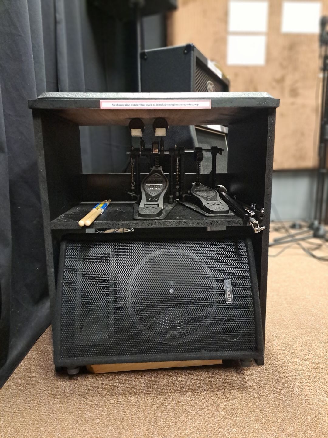 Czarny monitor perkusyjny Vonyx umieszczony w drewnianej obudowie z pedałami perkusyjnymi Perfect Sound i pałeczkami perkusyjnymi obok, na brązowym dywanie.