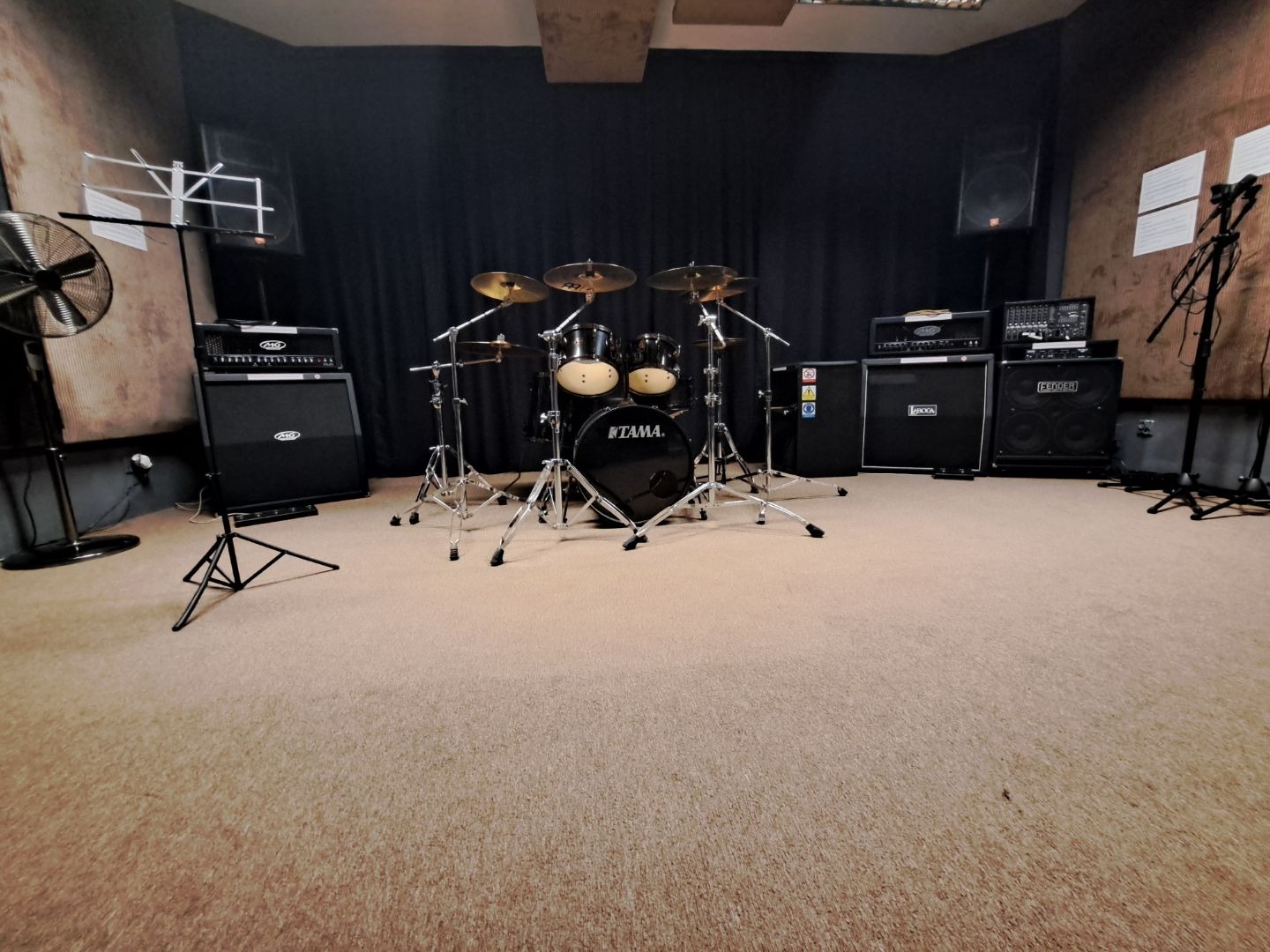 Wyposażona sala prób: perkusja Tama, wzmacniacze Peavey, Fender i inne, statyw na nuty, wentylator i mikrofony w pomieszczeniu z beżową wykładziną i czarną kotarą.