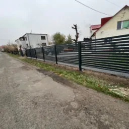 Nowoczesne, poziome, metalowe ogrodzenie panelowe w kolorze antracytowym, oddzielające posesję od drogi, z widocznymi domami w tle, sfotografowane w pochmurny dzień.