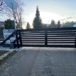 Taras Malena - Nowoczesna, przesuwna brama wjazdowa z poziomymi szczeblami, zamontowana na posesji z kostką brukową, widoczny napęd automatyczny i element ogrodzenia z imitacji kamienia.