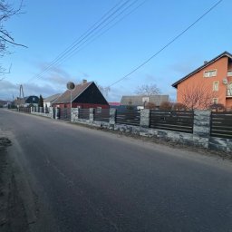 Taras Malena - Ulica z widokiem na posesje z ogrodzeniami panelowymi z elementami kamiennymi, w tle zabudowa jednorodzinna, pochmurne niebo.