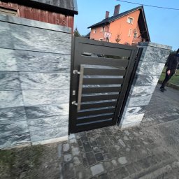 Taras Malena - Nowoczesna czarna brama z poziomymi szczeblami, zamontowana w ogrodzeniu z szarych, kamiennych bloków. W tle widoczny dom z pomarańczową elewacją i fragment chodnika z kostki brukowej, osoba...