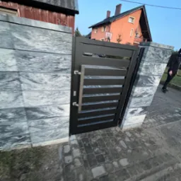 Nowoczesna czarna brama z poziomymi szczeblami, zamontowana w ogrodzeniu z szarych, kamiennych bloków. W tle widoczny dom z pomarańczową elewacją i fragment chodnika z kostki brukowej, osoba...