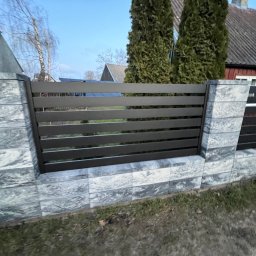 Taras Malena - Nowoczesne ogrodzenie z poziomych, grafitowych paneli między słupkami z szarej, imitującej kamień kostki, na tle domów i drzew